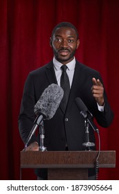 Vertical Portrait Of African-American Man Giving Speech Standing At Podium On Stage Against Red Curtain