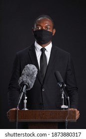 Vertical Portrait Of African-American Man Wearing Mask While Giving Speech Standing At Podium Against Black Background