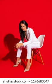 Vertical portrait of bad horny woman sitting on stool wearing white outfit looking at camera with angry sight wearing pump shoes isolated on vivid red background