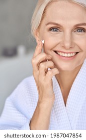 Vertical Portrait Of Beautiful Mid Age Older Adult 50 Years Old Blonde Woman Wears Bathrobe In Bathroom Applying Moisturizing Sunscreen Face Skin Care Cream Treatment, Looking At Camera.