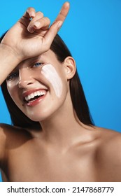 Vertical Portrait Of Beautiful Smiling Female Model With Lotion, Facial Cream Clay Mask On Face, Laughing And Looking Happy, Posing With Bare Glowing Shoulders After Shower, Blue Background