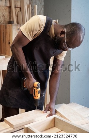 Vertical portrait of black carpenter using power drill while building wooden furniture in workshop