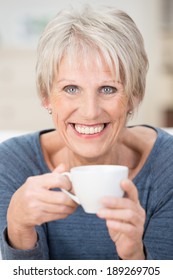 Vertical Portrait Of A Blond Short-haired Caucasian Senior Woman With Blue Eyes And A Big Smile Looking At Camera While Holding A White Ceramic Cup