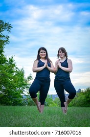 Vertical Portrait Of Brown Haired Women Doing Yoga Poses Outdoors. Blue Sky, Green Trees. Yogi Teachers Doing Standing Tree Pose, Modified And Mirroring Each Other, Hands In Prayer Position