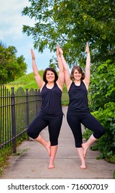 Vertical Portrait Of Brown Haired Women Doing Yoga Poses Outdoors. Blue Sky, Green Trees. Yogi Teachers Doing Standing Tree Pose, Modified And Mirroring Each Other. 