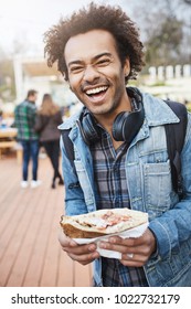Vertical portrait of charming unshaved dark-skinned guy holding tasty sandwich while walking with backpack in park or attending food festival, laughing out loud, expressing good mood