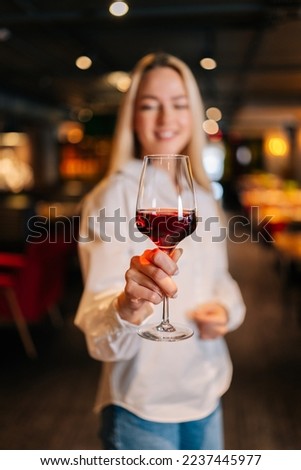 Vertical portrait of cheerful blonde woman holding glass of red wine standing in restaurant with luxury interior, looking away. Front view of cute female posing at cafe, blurred background.