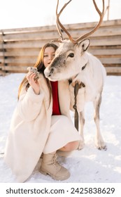 Retrato vertical de mujer joven alegre en abrigo de piel acariciando y alimentando lindo reno joven en la granja de ciervos nevados en el día soleado de invierno, sonriendo mirando al animal. Concepto de ecoturismo en invierno.