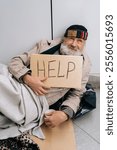 Vertical portrait of crying aged homeless man sitting on cold city sidewalk, holding sign asking for help. Sad elderly beggar male with face showing poverty and despair, alone in need of support.