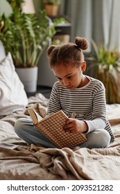 Vertical portrait of cute African-American girl reading book while sitting on bed in cozy interior