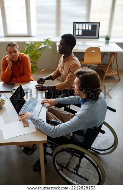 Vertical portrait of diverse business team\
discussing project at meeting table focus on young man using\
wheelchair in\
foreground
