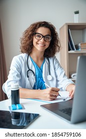 Vertical portrait of female doctor sitting on work desk and smiling at camera. Telemedicine, Medical online, e health concept. Doctor using laptop for work, video call video chat with colleagues.
