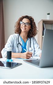 Vertical portrait of female doctor sitting on work desk and smiling. Telemedicine, Medical online, e health concept. Doctor using laptop for work, studying, video call and video chat with colleagues.