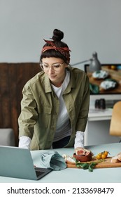 Vertical Portrait Of Female Food Photographer Using Laptop While Setting Up Scene In Studio