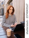 Vertical portrait of female freelancer with laptop sitting outdoor cafe, looking at camera. Focused redhead female using computer outdoor on cafe terrace. Lady using laptop for remote job.