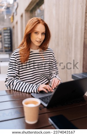 Vertical portrait of female freelancer with laptop sitting outdoor cafe, looking at camera. Focused redhead female using computer outdoor on cafe terrace. Lady using laptop for remote job.