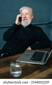 Vertical Portrait Of Gray-haired Mature Aged Business Man Talking On Mobile Phone Sitting At Table With Laptop Computer At Home Office. Serious Senior Older Male Having Conversation On Smartphone .