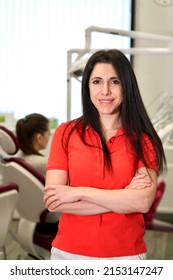 Vertical Portrait Of Happy Beautiful Female Doctor, Attractive Pretty Middle Aged Caucasian Armenian Ethnic Dentist In Red Polo Standing In Dental Office At Clinic, Smiling, Looking At Camera. 