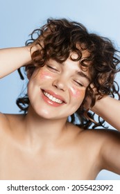 Vertical Portrait Of Happy Smiling Curly Girl With Glitter On Cheeks, Touching Her Hair, Washing Face And Body, Standing Against Blue Background