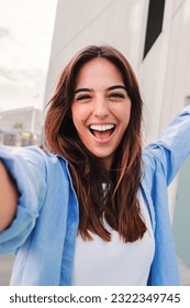 Vertical portrait of a happy young caucasian student lady looking at camera and taking a selfie having fun, standing outside. Laughing woman shooting a photo for social media at the university campus - Shutterstock ID 2322349745