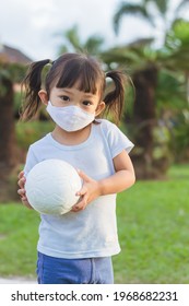 Vertical Portrait Image Of 2-3 Years Old Baby. Happy​ Playful Asian Child Girl Wearing Fabric Face Mask. She Play The Ball Toy At The Green Park Playground. Kids And Healthy.