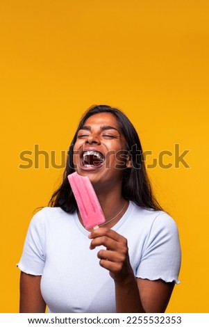 Vertical portrait of Indian woman laughing eating pink popsicle on yellow background. Asian woman eating ice cream.