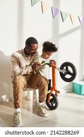 Vertical Portrait Of Loving Black Father Teaching Little Boy Riding Balance Bike At Home In Sunlight