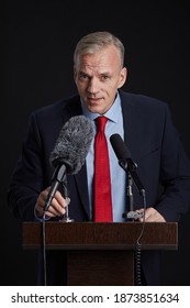 Vertical Portrait Of Mature Man Standing At Podium While Giving Speech To Microphone Against Black Background