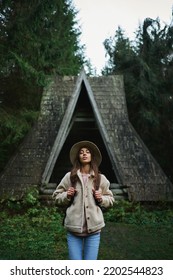 Vertical Portrait Millennial Backpacker Woman In Hat With Clothing Eyes Standing In Front Of Old Vintage Style Wooden Hut In The Dark Forest