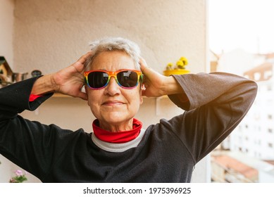 Vertical Portrait Of Older Lesbian Woman With Short Gray Hair Wearing Rainbow Pride Flag Sunglasses Smiling And Touching Her Head. Human Rights Activist. Lgbt Community.