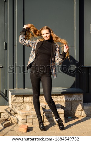 Vertical portrait of a pretty beautiful young girl with long hair with a smile in a park against the background of a wall of a business building. Photo taken in sunny weather outdoors in spring.