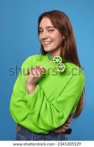 Vertical portrait of pretty cheerful girl wearing bright green sweater holding two lollipops in hand and smiling while looking at camera, isolated over blue background.