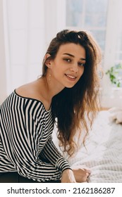 Vertical Portrait Of Pretty Photogenic Charming Girl With No Make-up, In Striped Blouse Baring Shoulders Sitting On Bed Over Day Light Coming Through Window. Natural Beauty Concept