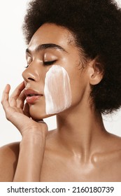 Vertical portrait of sensual black woman, gently touching her soft, healthy face with facial cream, skincare product on cheek, using lotion or daily care for better skin tone, pink background.