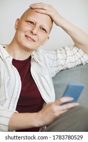 Vertical Portrait Of Smiling Bald Woman Rubbing Head And Looking Away Pensively While Sitting In Cozy Armchair At Home, Alopecia And Cancer Awareness