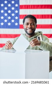 Vertical Portrait Of Smiling Black Man Putting Ballot In Bin Against American Flag In Background