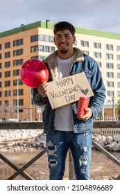 Vertical Portrait Of A Smiling Latino Man With A Happy Valentine's Day Sign,gift And Love Balloon Waiting For His Partner To Surprise Him And Celebrate Valentine's Day.