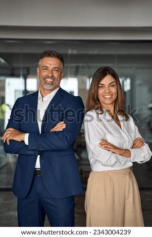 Vertical portrait of smiling mature Latin hispanic business man and European business woman standing arms crossed in office. Diverse colleagues, group team of confident professional business people.