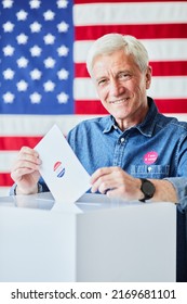 Vertical Portrait Of Smiling Senior Man Putting Ballot In Bin Against American Flag In Background