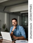 Vertical portrait of smiling young business man startup founder, happy Latin entrepreneur, confident business owner looking at camera using laptop computer technology working at corporate office desk.