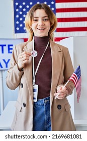 Vertical Portrait Of Smiling Young Woman Holding I Vote Sticker While Standing In Voting Station With American Flag