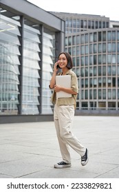 Vertical portrait of stylish asian girl student, talking on mobile phone while walking, holding laptop. - Shutterstock ID 2233829821
