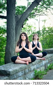 Vertical Portrait Of Two Brown Haired Women Doing Yoga Poses Outdoors In A Park. Green Trees And Sitting On Short Brick Wall. Modified Lotus Pose With Hands In Prayer Position. Yogi Instructors