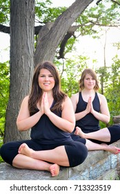 Vertical Portrait Of Two Brown Haired Women Doing Yoga Poses Outdoors In A Park. Green Trees And Sitting On Short Brick Wall. Modified Lotus Pose With Hands Prayer Position. Yogi Instructors Smiling.