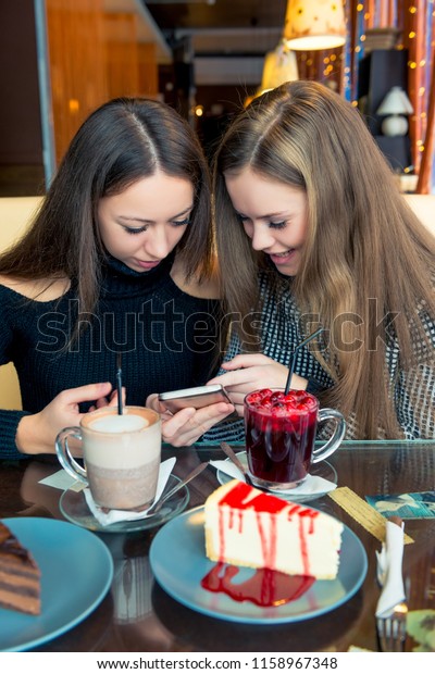 Vertical portrait of two girlfriends with a phone\
in the cafe are divided by\
secrets