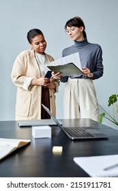 Vertical portrait of two young businesswomen reading document while standing in office against minimal grey background