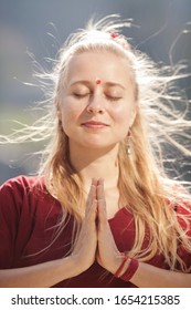 Vertical Portrait Of White Young Slim Woman With Long Blonde Hair, In Red T-shirt, With Earrings, Indian Mark On Forehead, Red String On Hand, Hands In Namaste, Eyes Closed, Outdoor On Sunny Day