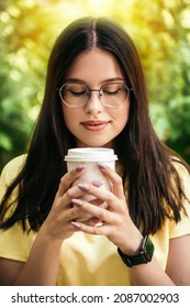 Vertical portrait woman smiling sitting park drink plastic white cup hot beverage takeaway coffee break, closed eyes smell aroma sniffs breathe notes caffeine wear glasses summer days copy space