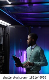 Vertical portrait of young African American data engineer holding laptop while working with supercomputer in server room lit by blue light, copy space