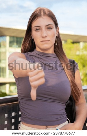 Vertical portrait Young angry caucasian woman pointing thumb down looking thoughtful outdoors. 